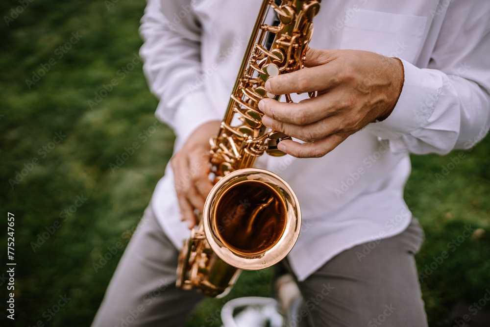 Naklejka premium Valmiera, Latvia - Augist 13, 2023 - Close-up of a person playing a golden saxophone, focusing on the hands and instrument against a blurred green background.