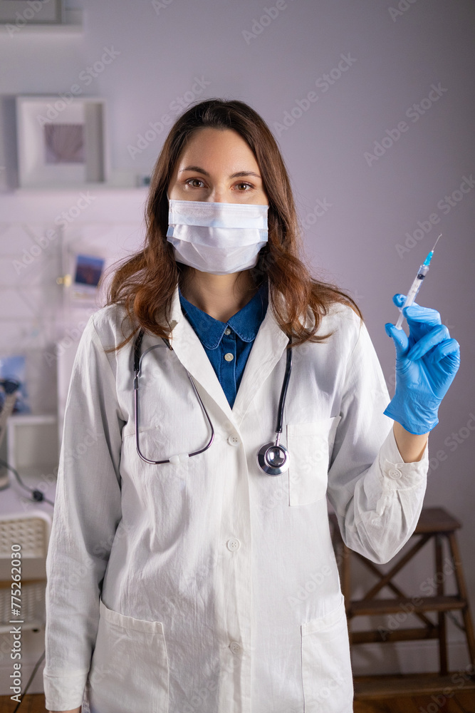 A young adult female doctor in a medical mask and white coat holds a syringe in her hands and looks at the camera A young adult female doctor in a medical mask and white coat holds a syringe in her hands and looks at the camera