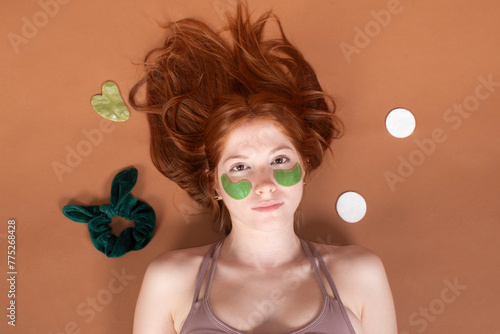 A redhead girl reclines with green eye patches on, surrounded by personal care items, against a brown background, creating a scene of tranquil self-care and relaxation.