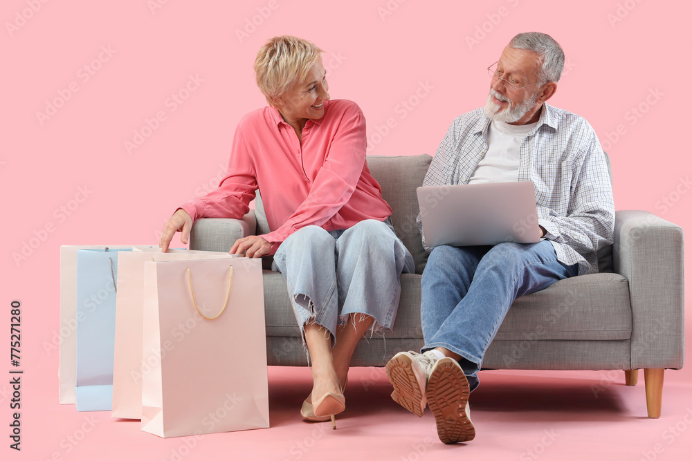 Mature couple with laptop and shopping bags sitting on sofa against pink background Mature couple with laptop and shopping bags sitting on sofa against pink background