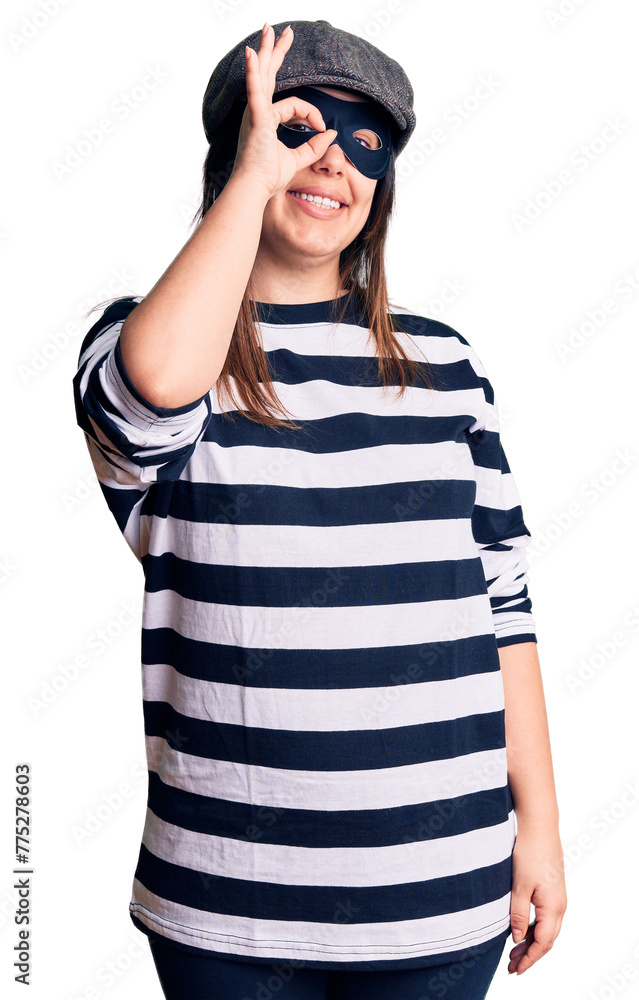 Young beautiful brunette woman wearing burglar mask doing ok gesture with hand smiling, eye looking through fingers with happy face. Young beautiful brunette woman wearing burglar mask doing ok gesture with hand smiling, eye looking through fingers with happy face.