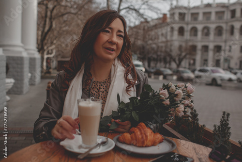 A charmig middle-aged woman drinks latte and eats  croissant in a french cafe on a city street. A woman with flowers is enjoing a wonderful and tasty breakfast photo