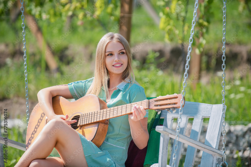 Young Woman sit on swing and playing guitar in summer park. Blonde Female model with guitar. Girl playing guitar outdoor. Singing Girl With Guitar. Summer music.