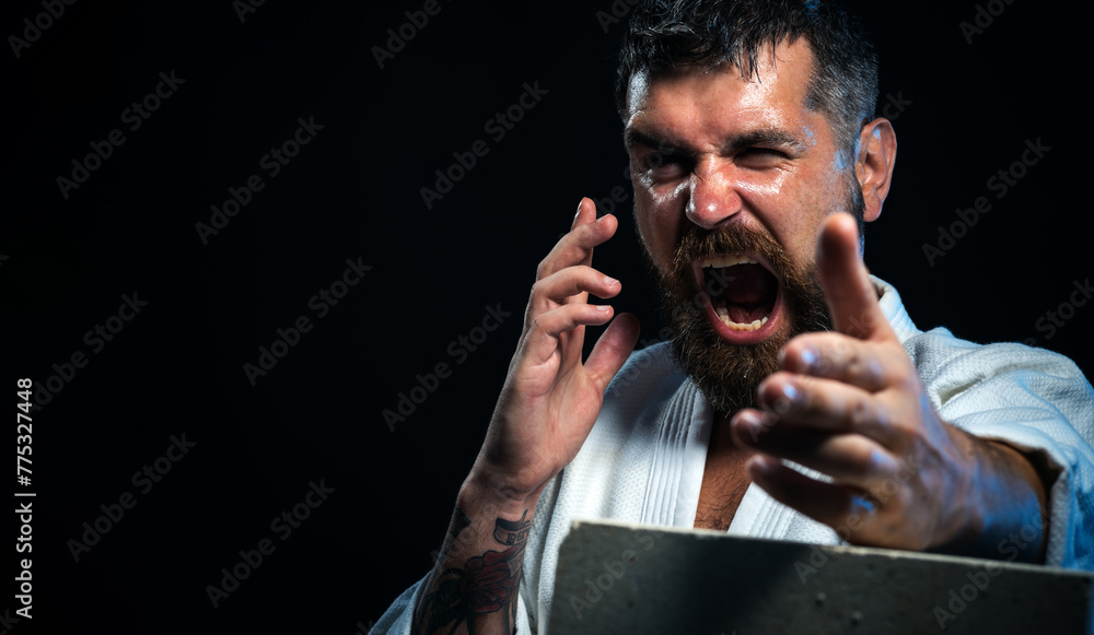 Bearded determined karate man breaking with hand concrete brick. Screaming karate fighter showing kick by hand. Angry karate master in kimono preparing for competition. Copy space for advertising. - obrazy, fototapety, plakaty 