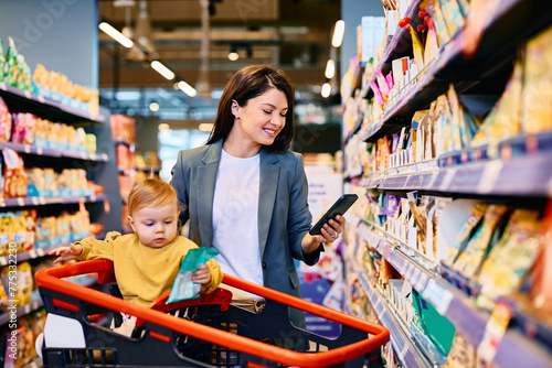 Happy woman using mobile phone app while buying with her small daughter at supermarket.