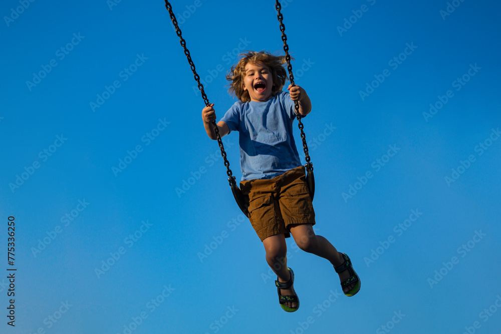 Little boy swinging on chain swing on city kids playground. Swing ride. Cute child having fun on a swing on summer sky background. Blonde little boy swings at kid playground. Child swinging high.