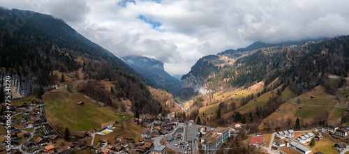 Aerial view of Murren, Switzerland, reveals its serene alpine charm. Chalets with sloping roofs along the main street merge into forested mountains. An overcast sky creates dynamic shadows
