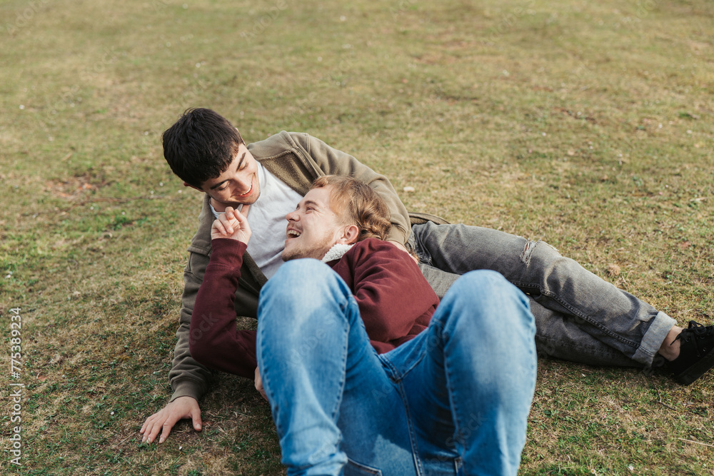 A carefree moment unfolds as one man reclines in the grass, sharing laughter and playful interaction with his partner outdoors A carefree moment unfolds as one man reclines in the grass, sharing laughter and playful interaction with his partner outdoors