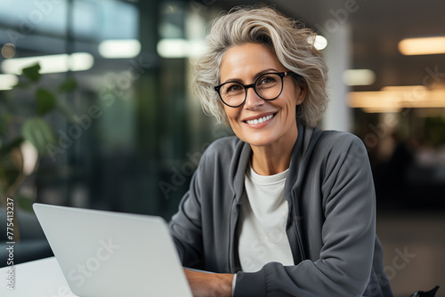 A mature businesswoman in her 50s with silver hair smiles while using her laptop in a casual cafe setting