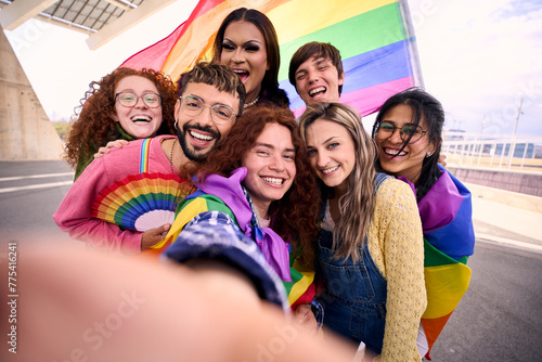 Group of young diverse friends taking a selfie looking at front camera of mobile celebrating gay pride day together holding rainbow flag. LGBT people community smiling and having fun outdoor 