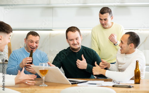 Group of men watching sports match online on laptop during friendly meeting with beer at home, excitedly celebrating winning score in game ..