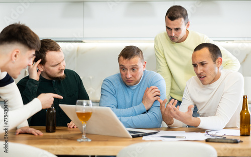 Concerned group of friends offering support to upset man receiving bad news, gathering together around laptop at home table during informal meeting with beer