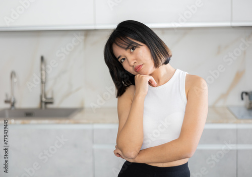 Disappointed young Asian lady standing by chairs in the kitchen with clean white cupboards
