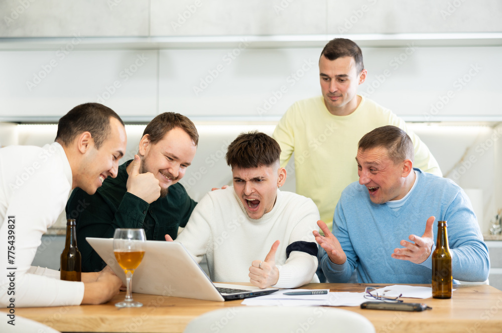Fototapeta premium Group of men watching sports match online on laptop during friendly meeting with beer at home, excitedly celebrating winning score in game ..