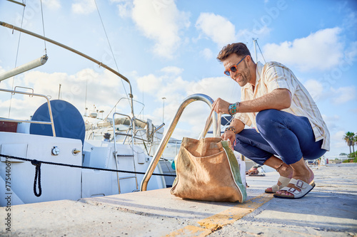 Photography 40s wealthy man ready to board the docked sailboat checks the bags with provisions at the pier of the yacht marina
