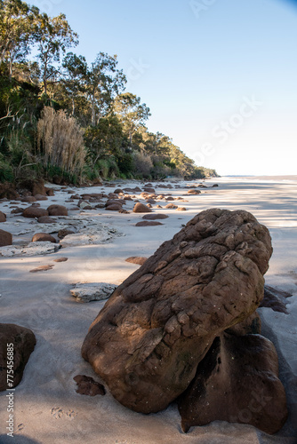 A large rock sits on a beach, surrounded by small rocks. The beach is rocky and the sky is clear photo