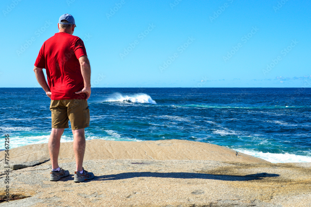 A senior male wearing a red shirt and beige shorts stands on the edge of a rocky cliff with his hands on his hips. The sky is bright blue. The ocean has white caps and waves rolling on the coastline. 