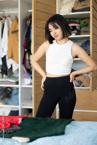 Distempered young Asian lady standing in front of wardrobe deciding which clothing to prefer