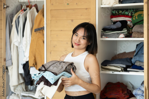 Glad young Asian woman posing by the wardrobe holding fold clothes in one hand photo