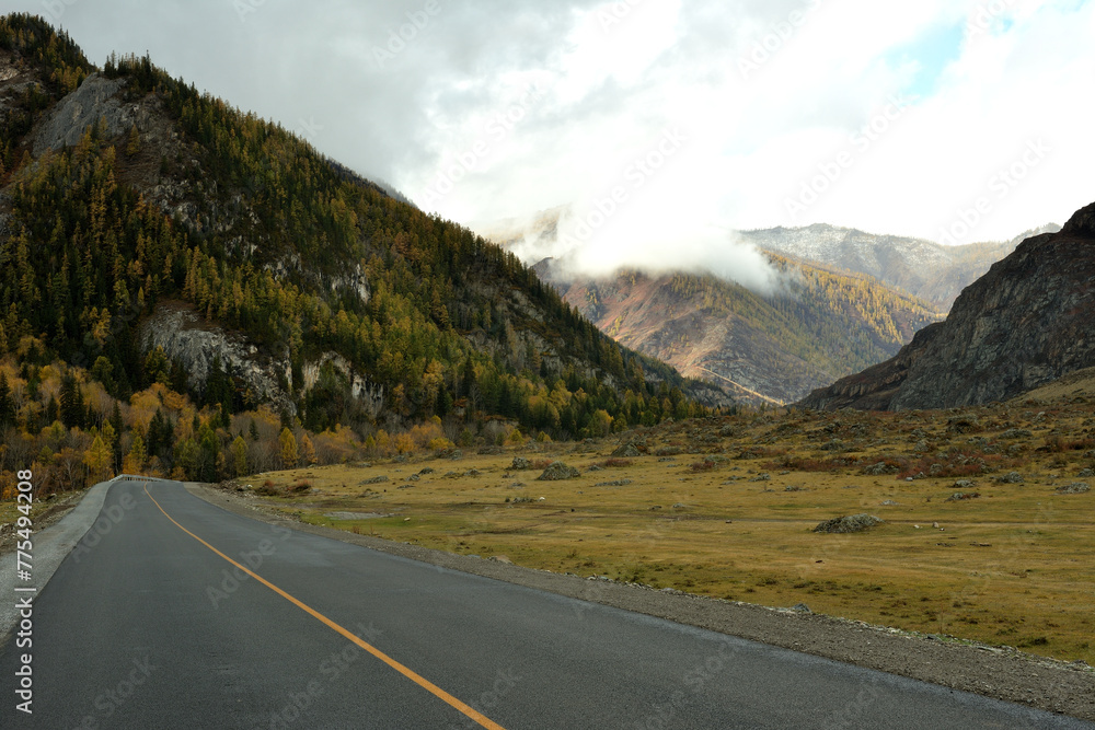 A two-lane asphalt road turns into a wide crevice towards high mountains with peaks in the clouds on a sunny autumn day.