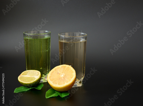 Two glasses of citrus juices on a black background, next to pieces of ripe lemon and lime with leaves.