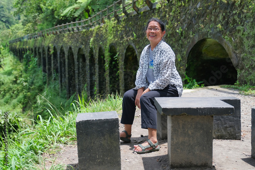 A woman sitting on a concrete bench with the pillars of an old brigde in the background; smiling, happy expression photo