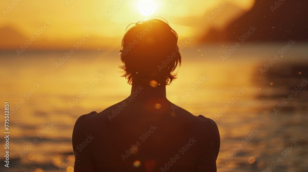 A man stands in front of a large body of water back to the camera as practices yoga using the tranquil atmosphere of the sunset . .