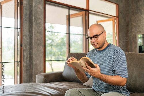 Asian man sitting reading book on cozey couch sofa in living room with sun light morning. People stay home part time relax at home. Good time quality life rest calm happiness