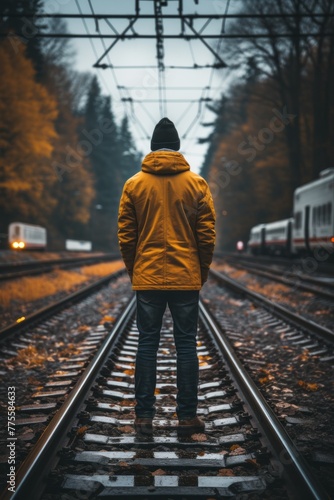 A man wearing a yellow jacket is standing on train tracks, appearing to be waiting or contemplating. The train tracks are clear with no train in sight