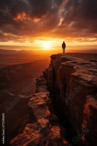 A man is standing on the edge of a cliff during sunset, overlooking the horizon as the sun sets in the background. The silhouette of the man is prominent against the colorful sky
