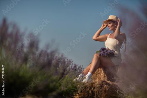 woman sitting in a field of lavender and wearing a straw hat. She is smiling and holding a bouquet of flowers. Scene is peaceful and serene, as the woman is surrounded by the beauty of nature