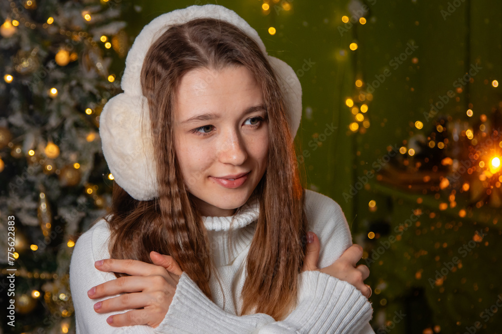 Close-up portrait of a young woman dressed in a white sweater and warm headphones posing against a background of golden bokeh and a Christmas tree. Close-up portrait of a young woman dressed in a white sweater and warm headphones posing against a background of golden bokeh and a Christmas tree.