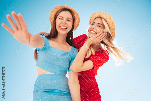 Two young beautiful smiling hipster woman in trendy summer same clothes. Carefree women in street in hats. Positive models at sunset. Cheerful and happy. Bottom view. They look at camera from above