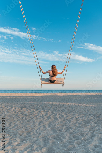 Young slim girl in swimsuit pushes on rope swing on blue sky, sea and beach background. Vertical frame.