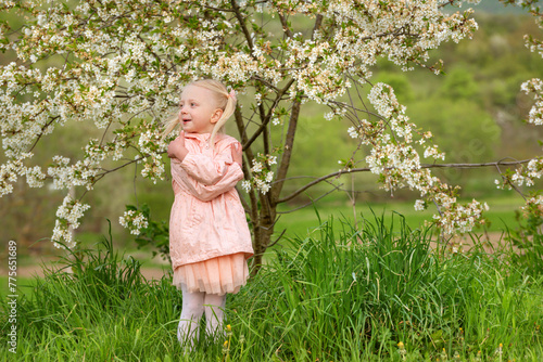 Pretty blonde girl in pink outfit walking in the garden next to blooming cherry tree in the sunny spring day.