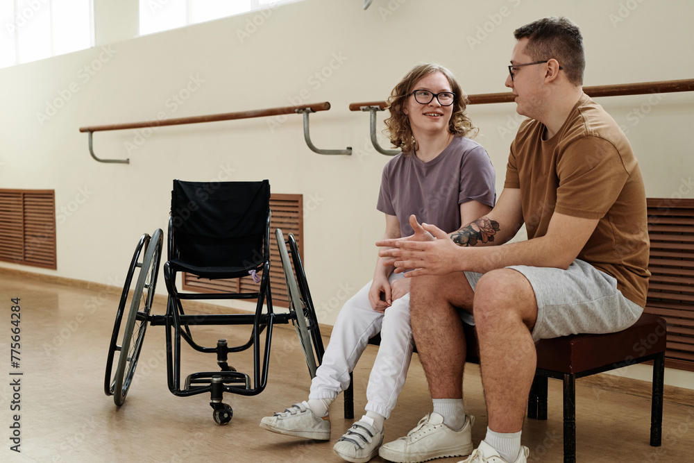 Fototapeta premium Long shot of modern Caucasian female wheelchair dancer and her partner sitting on bench in dance studio chatting during break
