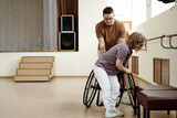 Long shot of young Caucasian man helping woman with disability to get into wheelchair in dance studio