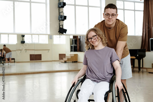 Medium long portarit of cheerful young woman in wheelchair and young man standing behind her in dance studio, copy space photo