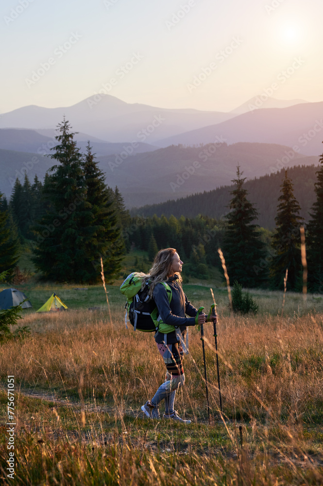 Woman hiker hiking outdoors at sunset. Sporty, slim woman traveling in mountains. Female tourist carrying backpack, using trekking sticks, admiring beautiful landscape. Concept of harmony with nature.