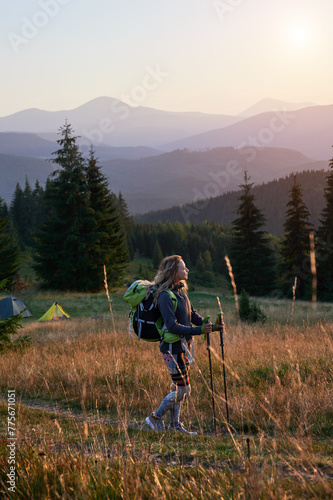 Woman hiker hiking outdoors at sunset. Sporty, slim woman traveling in mountains. Female tourist carrying backpack, using trekking sticks, admiring beautiful landscape. Concept of harmony with nature.