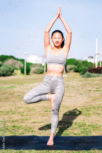 young asian woman in sportswear doing tree position on her yoga mat at park, active and healthy lifestyle concept