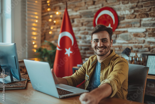 Happy young guy sitting at desk with flag of Turkey, using laptop, showing thumb up gesture at home  photo