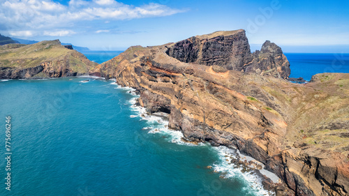 A steep, rocky coastline with light blue ocean water at its base. Visible are green grassy areas and paths along the cliffs. It's the beauty of nature in Madeira, the PR8 trail. © Sebastian