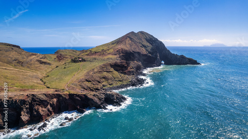 A steep, rocky coastline with light blue ocean water at its base. Visible are green grassy areas and paths along the cliffs. It's the beauty of nature in Madeira, the PR8 trail.