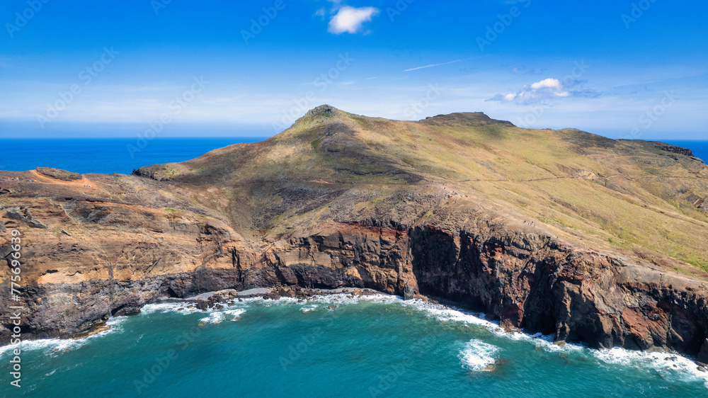 The rocky coastline with a light blue sea, rocks, and green hills. It's a sunny day. PR8 trail in Madeira. It looks like a beautiful coastal landscape.