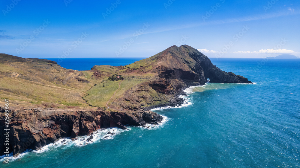 Fototapeta premium A steep, rocky coastline with light blue ocean water at its base. Visible are green grassy areas and paths along the cliffs. It's the beauty of nature in Madeira, the PR8 trail.