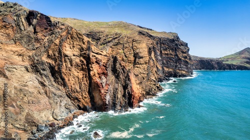 A steep, rocky coastline with light blue ocean water at its base. Visible are green grassy areas and paths along the cliffs. It's the beauty of nature in Madeira, the PR8 trail.