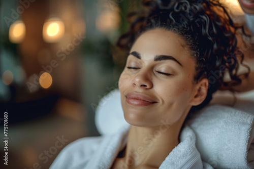 A close-up portrait of an attractive woman receiving a facial massage at a spa salon, smiling and relaxing with closed eyes, wearing a white robe. The focus is on her face showing tranquility and rela