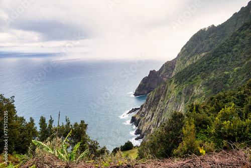 The photo depicts a steep, green mountainside in Madeira, sloping down to the blue sea. Waves crash against the rocky coast, and dense clouds dominate the scene, creating an image of untouched nature.