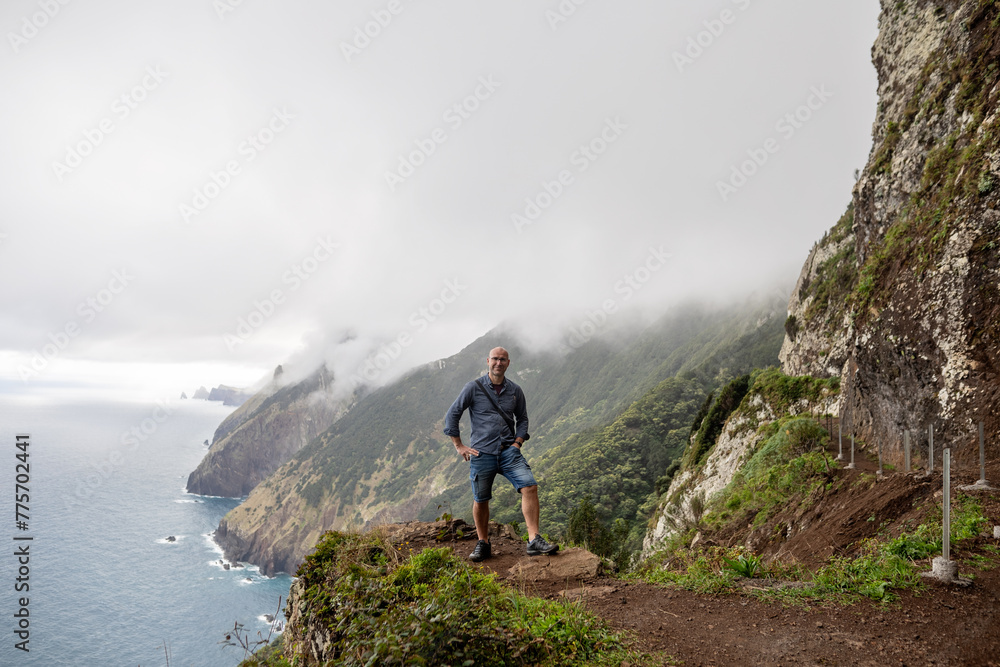 The picture shows a person standing at the edge of a cliff, overlooking the ocean and the mountains of Madeira. The mountains are steep, covered in greenery, and partially shrouded in mist. The picture shows a person standing at the edge of a cliff, overlooking the ocean and the mountains of Madeira. The mountains are steep, covered in greenery, and partially shrouded in mist.